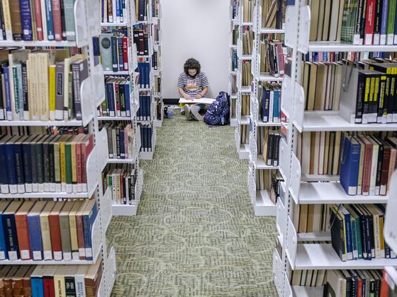 A student sits on the floor in Hoover Library between the aisle of book shelves.