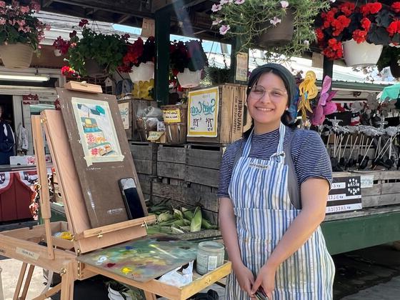 Sarah Mendez at an easel wearing an apron. Behind her is a market stand with flowers.