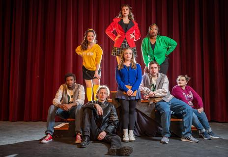 The full cast of Heathers The Musical. They are in vivid colored clothing with sad expressions.
